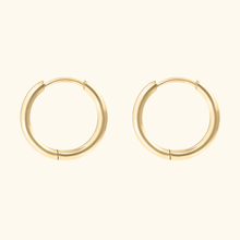 Load image into Gallery viewer, Gold-colored huggie hoop earrings on a light yellow background.