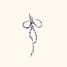 Load image into Gallery viewer, silver colored bow charm with a light yellow background.
